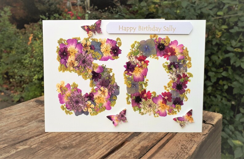 May include: A handmade birthday card with the number 50 formed from dried flowers in shades of purple, pink, and yellow. The card includes the text "Happy Birthday Sally" and decorative butterfly accents.