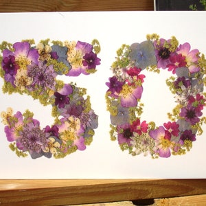 May include: A white card features the number "50" formed from dried flowers. The flowers are primarily purple, with accents of pink, yellow, and green. The card is set against a wooden surface.