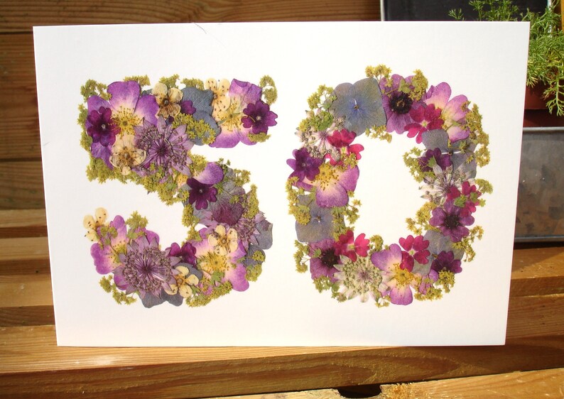 May include: A white card features the number "50" formed from dried flowers. The flowers are primarily purple, with accents of pink, yellow, and green. The card is set against a wooden surface.