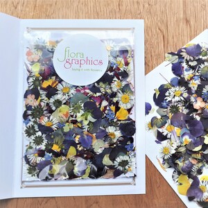 May include: A clear plastic bag filled with assorted pressed dried flowers in shades of blue, purple, yellow, and white, featuring a circular white label with the text flora graphics Saying it with flowers, placed on a white card.