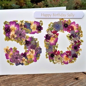 May include: A white birthday card with the number "50" formed from purple, pink, and yellow dried flowers. The card includes a banner that reads "Happy Birthday Sally".