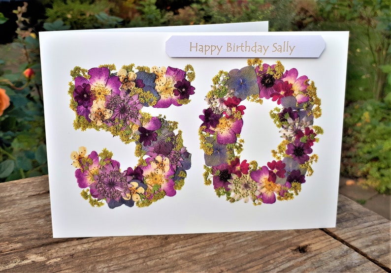 May include: A white birthday card with the number "50" formed from purple, pink, and yellow dried flowers. The card includes a banner that reads "Happy Birthday Sally".