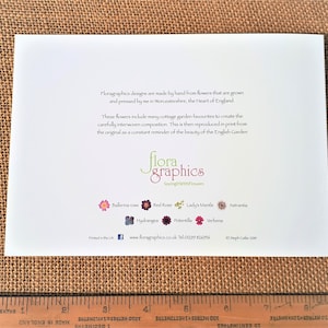May include: A white card with text and floral illustrations, featuring the words "flora graphics" and "SayingWithFlowers." The card includes a ruler at the bottom and is set against a textured, brown background.