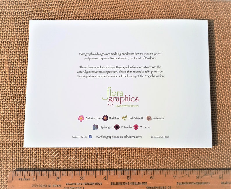 May include: A white card with text and floral illustrations, featuring the words "flora graphics" and "SayingWithFlowers." The card includes a ruler at the bottom and is set against a textured, brown background.