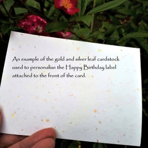 May include: A white cardstock with gold and silver leaf flecks. The card has text that reads, "An example of the gold and silver leaf cardstock used to personalise the Happy Birthday label attached to the front of the card."