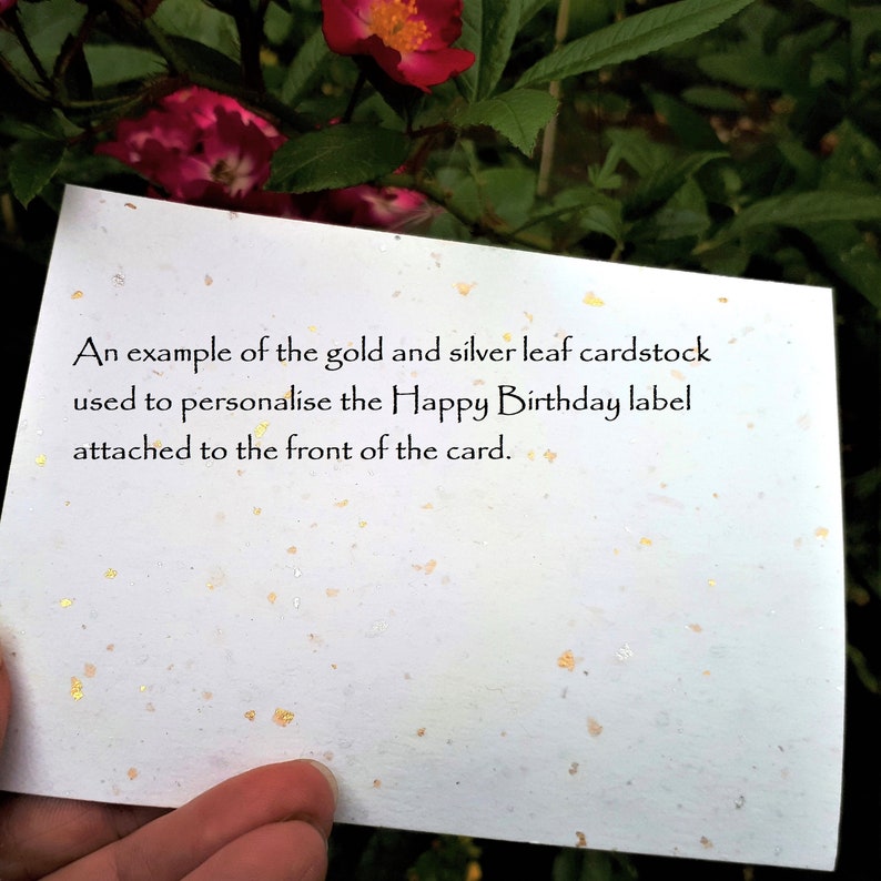 May include: A white cardstock with gold and silver leaf flecks. The card has text that reads, "An example of the gold and silver leaf cardstock used to personalise the Happy Birthday label attached to the front of the card."