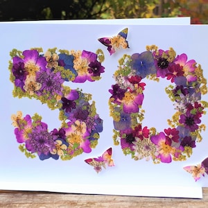 May include: A white greeting card with the number "50" formed from purple, pink, and yellow pressed flowers. Three butterfly accents are scattered around the numbers. The card is on a wooden surface.