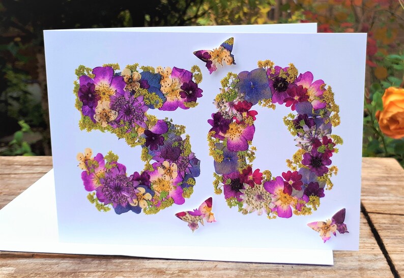 May include: A white greeting card with the number "50" formed from purple, pink, and yellow pressed flowers. Three butterfly accents are scattered around the numbers. The card is on a wooden surface.