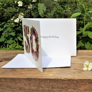 May include: A white birthday card with the number "50" formed by colorful flowers. The card is open, revealing the words "Happy Birthday" inside. The card is on a wooden surface with greenery in the background.