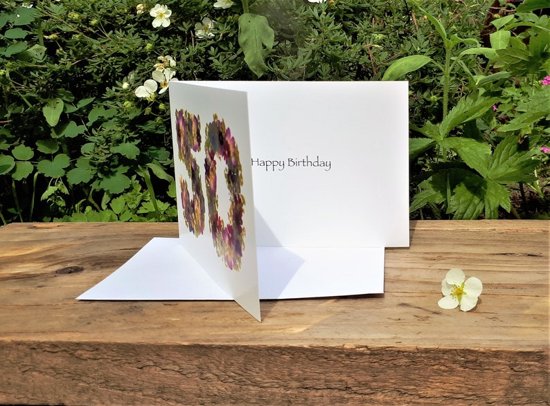 May include: A white birthday card with the number "50" formed by colorful flowers. The card is open, revealing the words "Happy Birthday" inside. The card is on a wooden surface with greenery in the background.