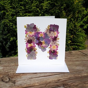 May include: A square white greeting card featuring the letter M formed by an arrangement of pressed dried flowers in shades of purple, pink, and yellow. The floral design is centered on the front of the card, creating a botanical letter.