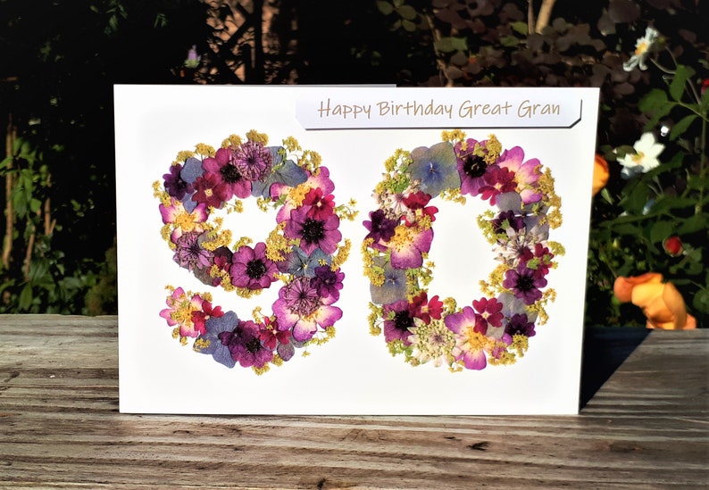 May include: A white card with the words "Happy Birthday Great Gran" printed on it. The number 90 is made from pressed flowers in shades of purple, pink, blue, and yellow.