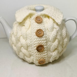 May include: A white ceramic teapot covered by a cream-colored knitted tea cozy. The cozy features a cable knit pattern, a folded collar, and four light brown wooden buttons down the center, resembling a cardigan sweater design.