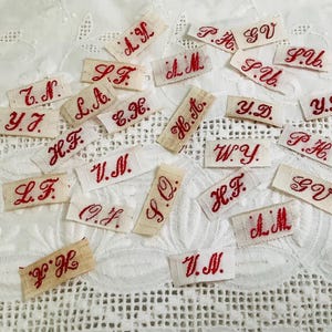 May include: A collection of small, rectangular fabric labels in off-white, each featuring embroidered red initials in a cursive script. The labels include various letter combinations such as A.K, L.F, Y.J, H.F, V.N, O.F, S.Q, H.T, W.Y, P.H, G.V, S.U, and Y.D.