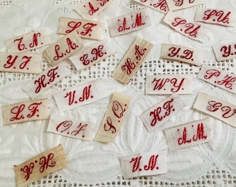 May include: A collection of small, rectangular fabric labels in off-white, each featuring embroidered red initials in a cursive script. The labels include various letter combinations such as A.K, L.F, Y.J, H.F, V.N, O.F, S.Q, H.T, W.Y, P.H, G.V, S.U, and Y.D.
