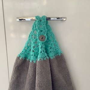 May include: A grey kitchen towel with a teal crocheted top and a wooden button. The towel is hanging on a silver hook.