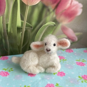 May include: A small, white, felted lamb figurine with pink ears, lying on a blue and white floral background.