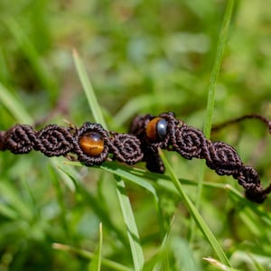 Puede incluir: Dos pulseras de macramé con cordón marrón y cuentas de ojo de tigre descansan sobre hierba verde. Las pulseras tienen un diseño tejido con una cuenta central. El fondo es verde borroso.