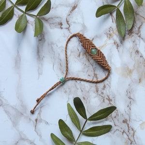Op de afbeelding: Een handgemaakte macramé armband met een groene edelsteen in het midden. De armband is gemaakt van bruin koord en heeft een verstelbare sluiting. De achtergrond is een marmerpatroon met groene bladeren.