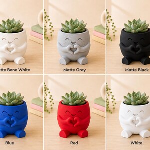 May include: A collection of six small, matte-finish ceramic planters, each holding a succulent plant. The planters are in various colors: bone white, gray, black, blue, red, and white. Each planter has a smiling face design with hands forming a heart shape.