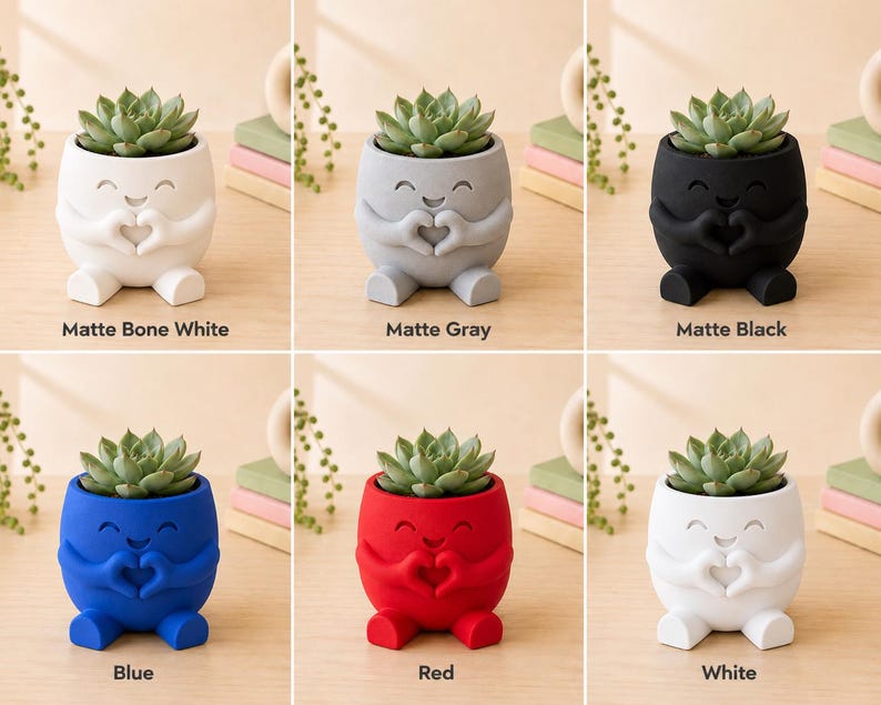 May include: A collection of six small, matte-finish ceramic planters, each holding a succulent plant. The planters are in various colors: bone white, gray, black, blue, red, and white. Each planter has a smiling face design with hands forming a heart shape.
