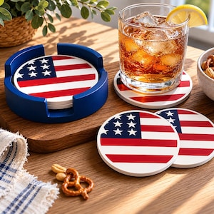 May include: A set of drink coasters featuring the American flag design. The coasters have a white base with red and white stripes and a blue field with white stars. A blue coaster holder is also included.