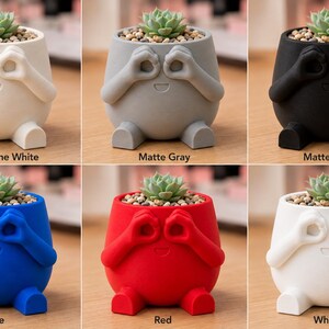 May include: A collection of whimsical, small succulent planters in matte finishes: bone white, gray, black, blue, red, and white. Each planter has a round body with arms and legs, and a face with hands over the eyes. The planters are filled with soil and a small succulent.