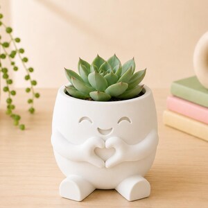 May include: A white ceramic planter shaped like a smiling character, with arms forming a heart shape. The planter holds a green succulent plant. The planter has a cute design and is sitting on a wooden surface.