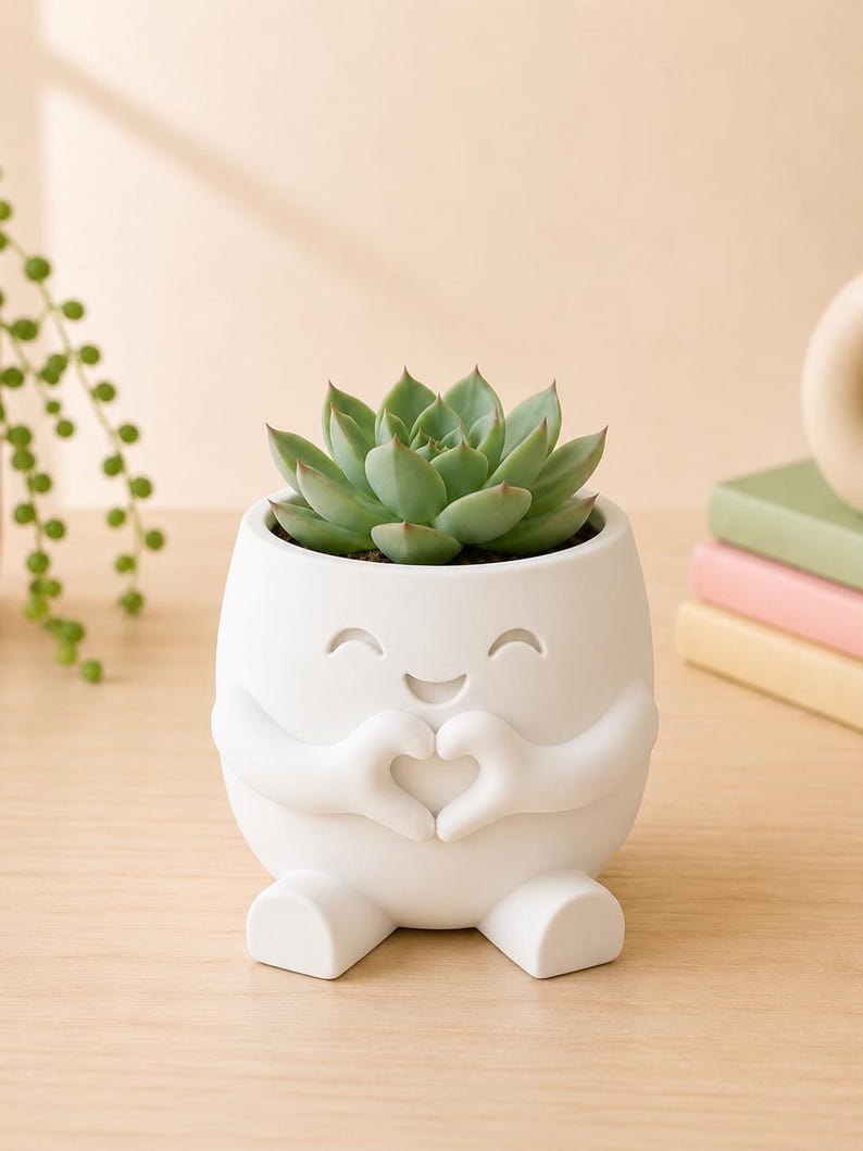 May include: A white ceramic planter shaped like a smiling character, with arms forming a heart shape. The planter holds a green succulent plant. The planter has a cute design and is sitting on a wooden surface.