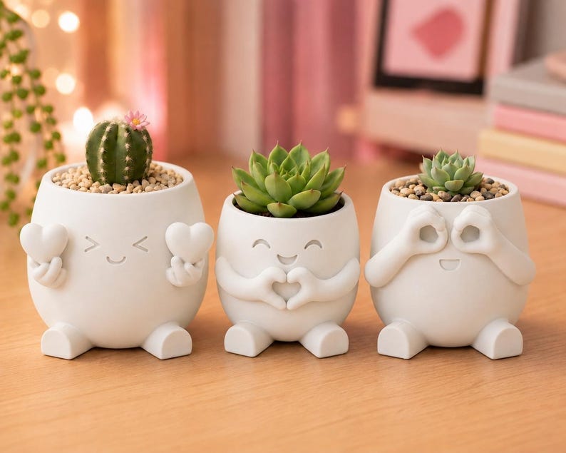 May include: Three white ceramic planters with smiling faces and small feet. One holds a cactus with a pink flower, another a green succulent, and the third a small succulent. The planters have arms that form hearts and the 'okay' symbol.