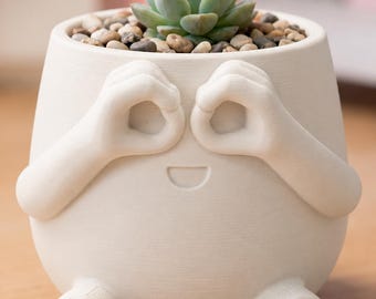 May include: A whimsical off-white ceramic planter with a smiling face design. The planter features arms forming eye-like circles and small feet. It is filled with small pebbles and a green succulent plant.