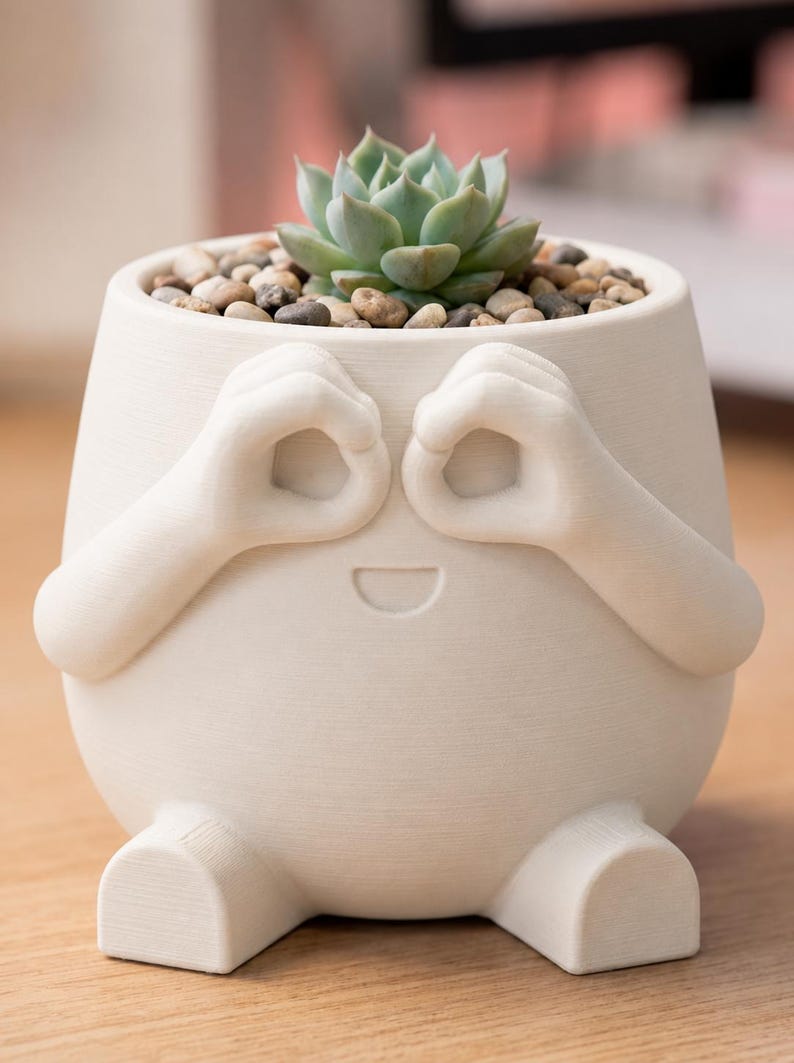 May include: A whimsical off-white ceramic planter with a smiling face design. The planter features arms forming eye-like circles and small feet. It is filled with small pebbles and a green succulent plant.