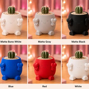 May include: A collection of small, colorful planters, each shaped like a character with a smiling face and heart-shaped hands. Each planter holds a small cactus. Colors include matte bone white, gray, black, blue, red, and white. Text on the image reads "Matte Bone White", "Matte Gray", "Matte Black", "Blue", "Red", and "White".