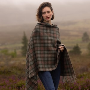 Könnte beinhalten: Eine Frau trägt einen braunen, grünen und grauen karierten Poncho mit Fransen. Sie trägt blaue Jeans und braune Stiefel. Der Hintergrund zeigt ein Feld mit lila Blumen und eine neblige Landschaft.