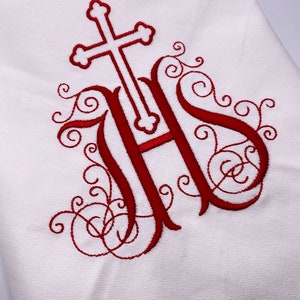Könnte beinhalten: Weißer Stoff mit einem roten Stickmuster. Das Design zeigt ein Kreuz über den Buchstaben IHS mit dekorativen Schnörkeln. Der Stoff ist gefaltet und zeigt eine strukturierte Oberfläche. Die Stickerei ist detailliert und die Farbe ist ein tiefes Rot.