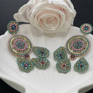 Könnte beinhalten: Ein Paar verzierte, perlenbesetzte Ohrringe. Die Ohrringe haben einen kreisförmigen oberen Teil mit einem floralen Design, verbunden mit tropfenförmigen Anhängern. Die Farbpalette umfasst türkisfarbene, goldene, rosa und violette Perlen.