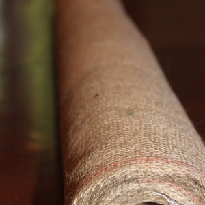 May include: Rolled-up natural fiber rug, showcasing a close-up view of its woven texture. The rug is a warm, neutral tan color with subtle red stitching along the edges. The roll reveals a cardboard core.