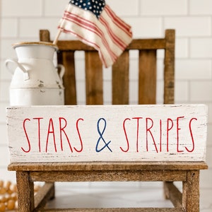 Puede incluir: Un letrero rectangular de madera blanca con bordes desgastados que presenta el texto en rojo y azul STARS & STRIPES. El letrero descansa sobre un peque&ntilde;o taburete de madera marr&oacute;n, colocado frente a una jarra de metal blanca y una bandera estadounidense en miniatura.
