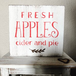 May include: A white wooden block sign with red lettering that reads "FRESH APPLES cider and pie." The sign has a distressed, rustic look and a small brown leaf design. The sign is displayed on a small, white, wooden table.