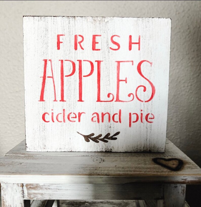 May include: A white wooden block sign with red lettering that reads "FRESH APPLES cider and pie." The sign has a distressed, rustic look and a small brown leaf design. The sign is displayed on a small, white, wooden table.