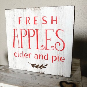 May include: A white wooden sign with the words "FRESH APPLES cider and pie" in red lettering. The sign has a distressed, rustic look, with a small leaf design below the text. The sign is square and sits on a wooden surface.