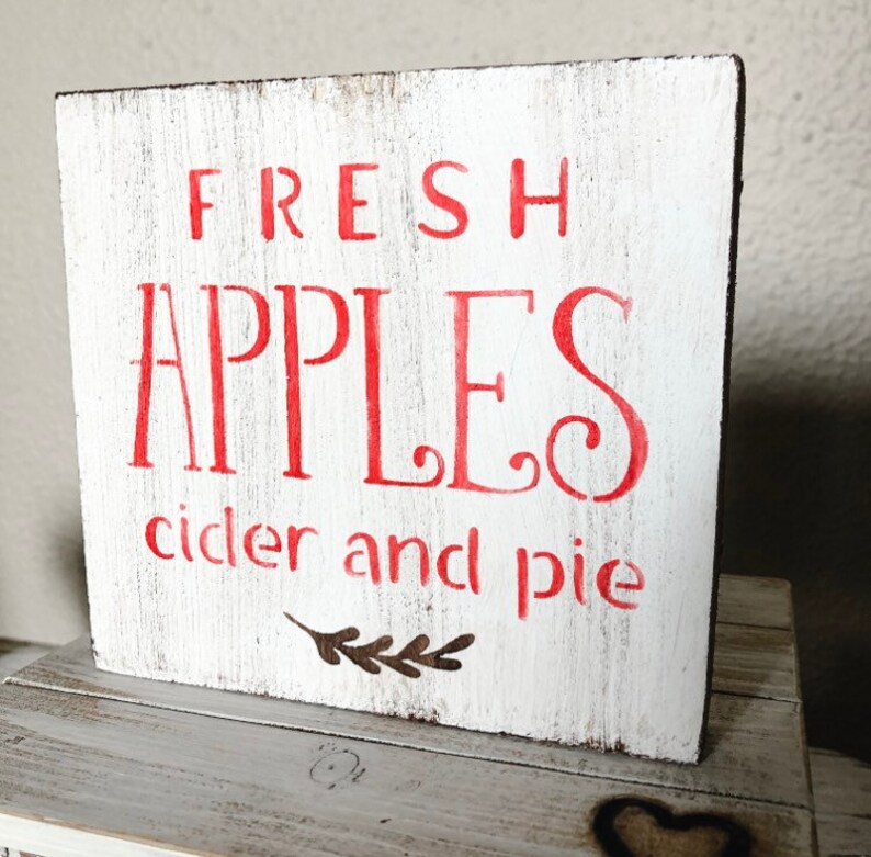 May include: A white wooden sign with the words "FRESH APPLES cider and pie" in red lettering. The sign has a distressed, rustic look, with a small leaf design below the text. The sign is square and sits on a wooden surface.