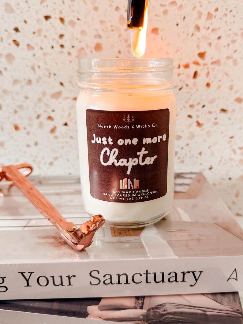 K&ouml;nnte beinhalten: Eine brennende Sojawachskerze in einem Glas mit einem braunen Etikett mit der Aufschrift "Just one more chapter". Ein kupferfarbener Dochtschneider liegt auf einem Stapel B&uuml;cher. Die Kerze ist 198 g.