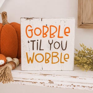 May include: A square white wooden sign with a distressed finish featuring the orange, brown, and yellow text GOBBLE 'TIL YOU WOBBLE. The sign is positioned next to a textured orange fabric pumpkin and a small decorative tassel with wooden beads.