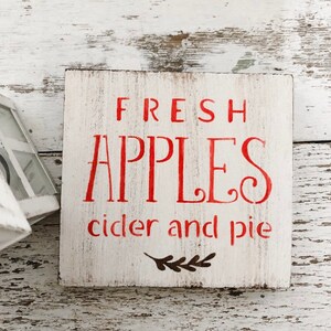 May include: A square, white wooden sign with red lettering that reads "FRESH APPLES cider and pie." The sign has a distressed, rustic look, with a small sprig graphic at the bottom. Perfect for kitchen or dining room decor.
