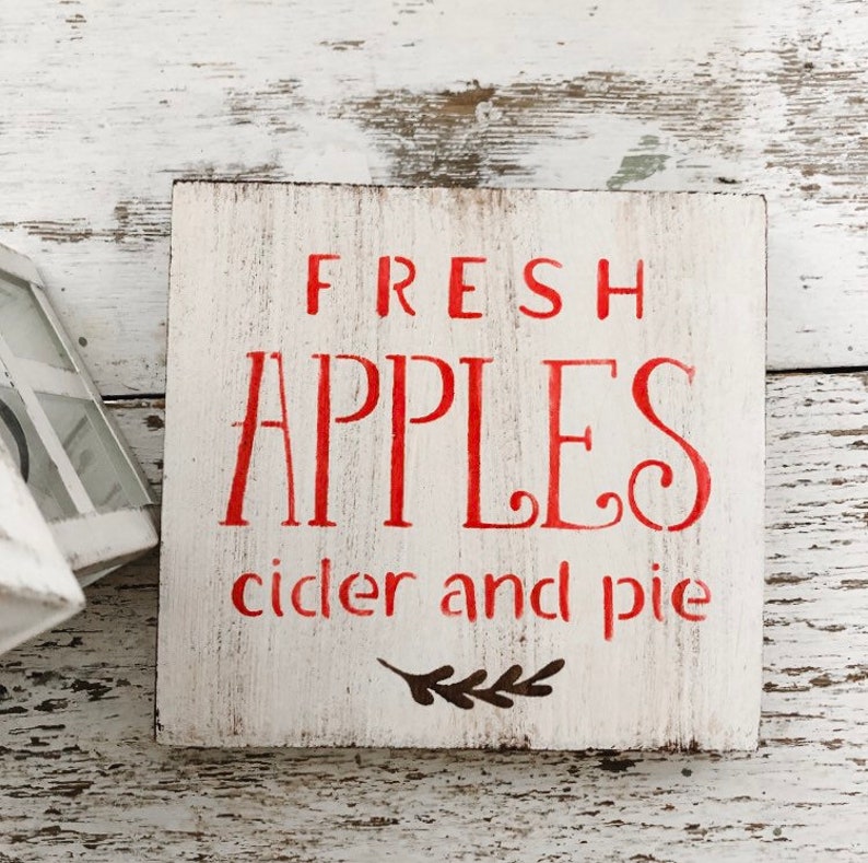 May include: A square, white wooden sign with red lettering that reads "FRESH APPLES cider and pie." The sign has a distressed, rustic look, with a small sprig graphic at the bottom. Perfect for kitchen or dining room decor.