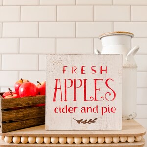 May include: A rustic wooden sign with the words "FRESH APPLES cider and pie" in red lettering. A wooden crate filled with red apples sits to the left. A white milk jug is in the background. The sign is on a wooden tray.