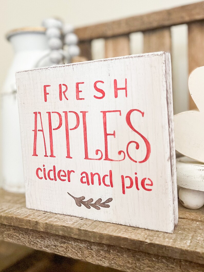 May include: A rustic, white wooden sign with red lettering that reads "FRESH APPLES cider and pie." The sign has a distressed finish and a small leaf design. It is displayed on a wooden shelf, suggesting a fall or harvest theme.