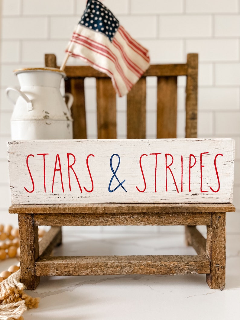Puede incluir: Un letrero rectangular de madera blanca con bordes desgastados que presenta el texto en rojo y azul STARS & STRIPES. El letrero descansa sobre un peque&ntilde;o taburete de madera marr&oacute;n, colocado frente a una jarra de metal blanca y una bandera estadounidense en miniatura.