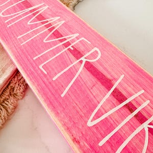 May include: A rectangular wooden sign painted in a vibrant hot pink colour with a visible wood grain texture. The text "SUMMER VIBES" is written across the surface in a white, hand-lettered script font. The sign has a flat, elongated shape.